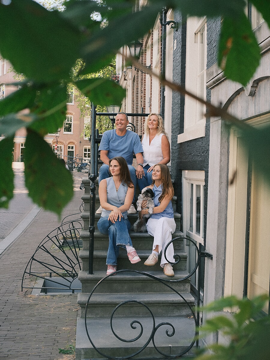 Family of four and their small dog sitting on the stone steps of a canal house in Amsterdam, framed through green leaves