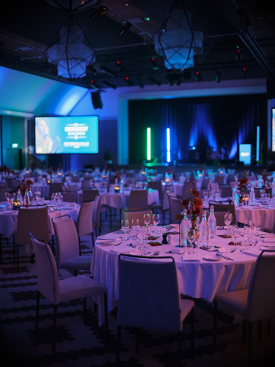 Gala ballroom before the guests arrive, round tables in white linen under blue and green stage light, an Amsterdam business event