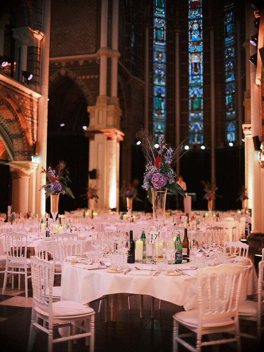 Gala dinner inside a converted gothic church, white chairs and tall floral centrepieces below stained glass, a business gala in Amsterdam
