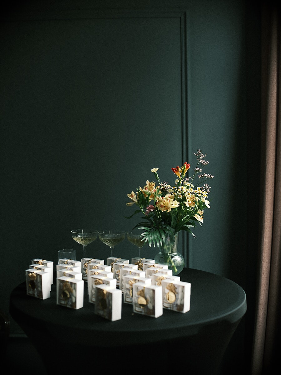 Rows of small white gift boxes with gold charms on a dark table beside coupe glasses and flowers, product photography for an Amsterdam business