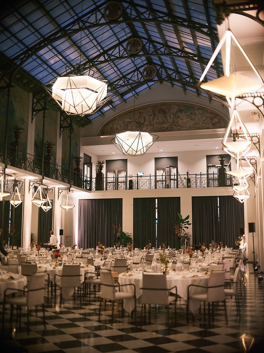 Grand hall with a glass barrel roof and painted coats of arms, round tables set beneath geometric pendant lamps in Amsterdam