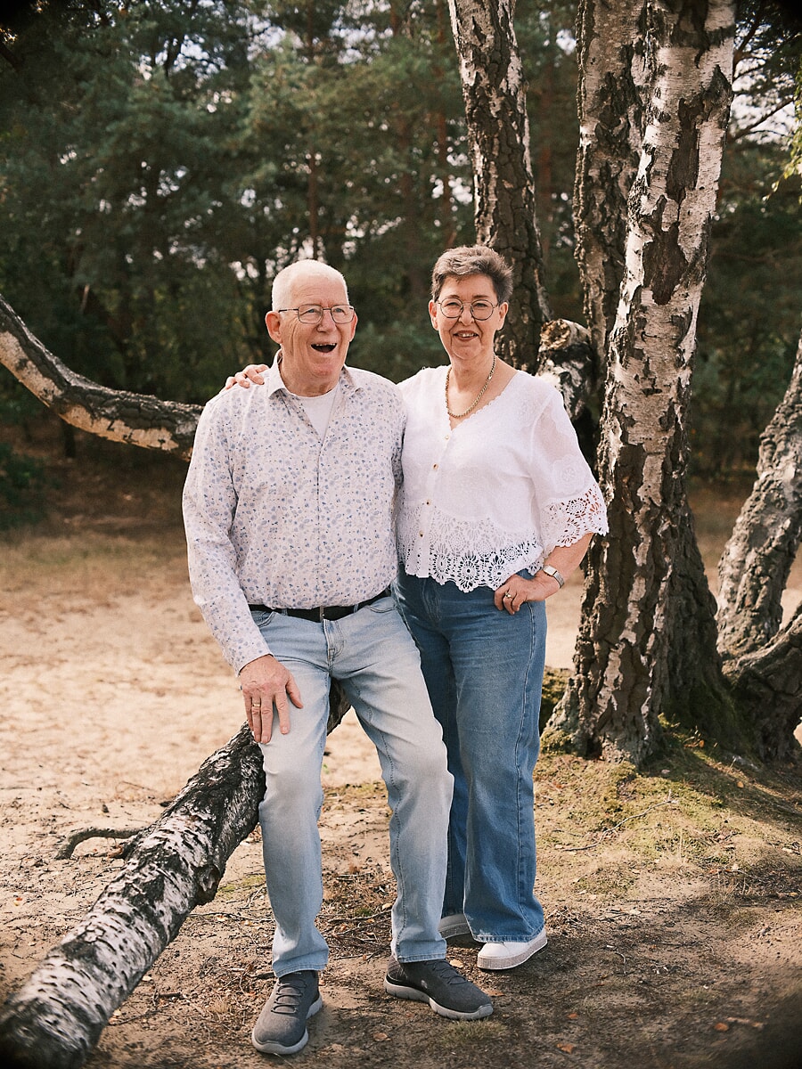 Grandparents arm in arm, laughing beside a leaning birch trunk in sunlit woodland