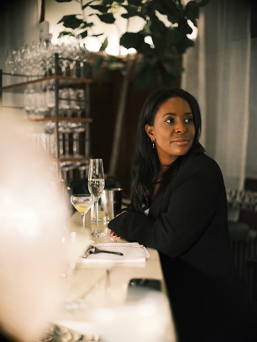 Guest in a black blazer leaning on a marble bar with a champagne flute, a fig tree behind her, a business reception in Amsterdam