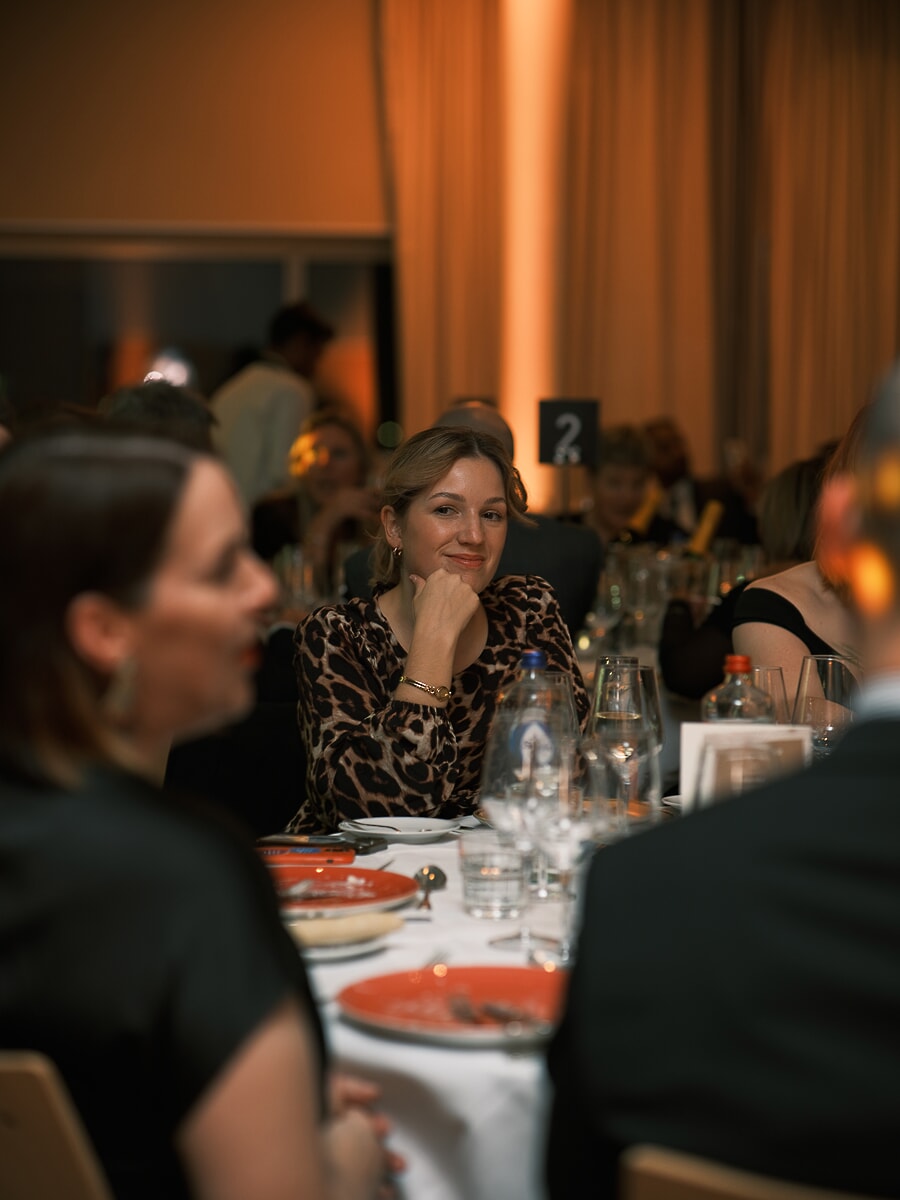 Guest in leopard print resting her chin on her hand at a dinner table, warm uplight behind her, a business dinner in Amsterdam