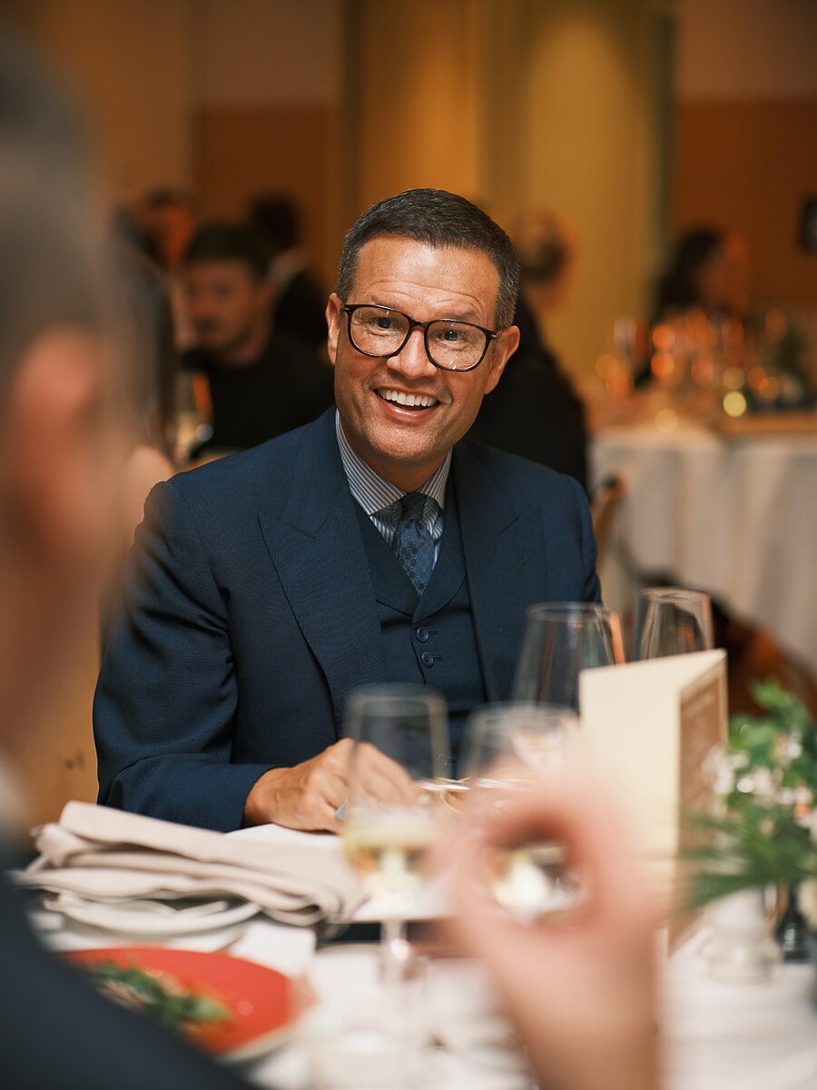 Guest in a navy three-piece suit laughing at a dinner table, wine glasses catching the light in front of him, a business dinner in Amsterdam