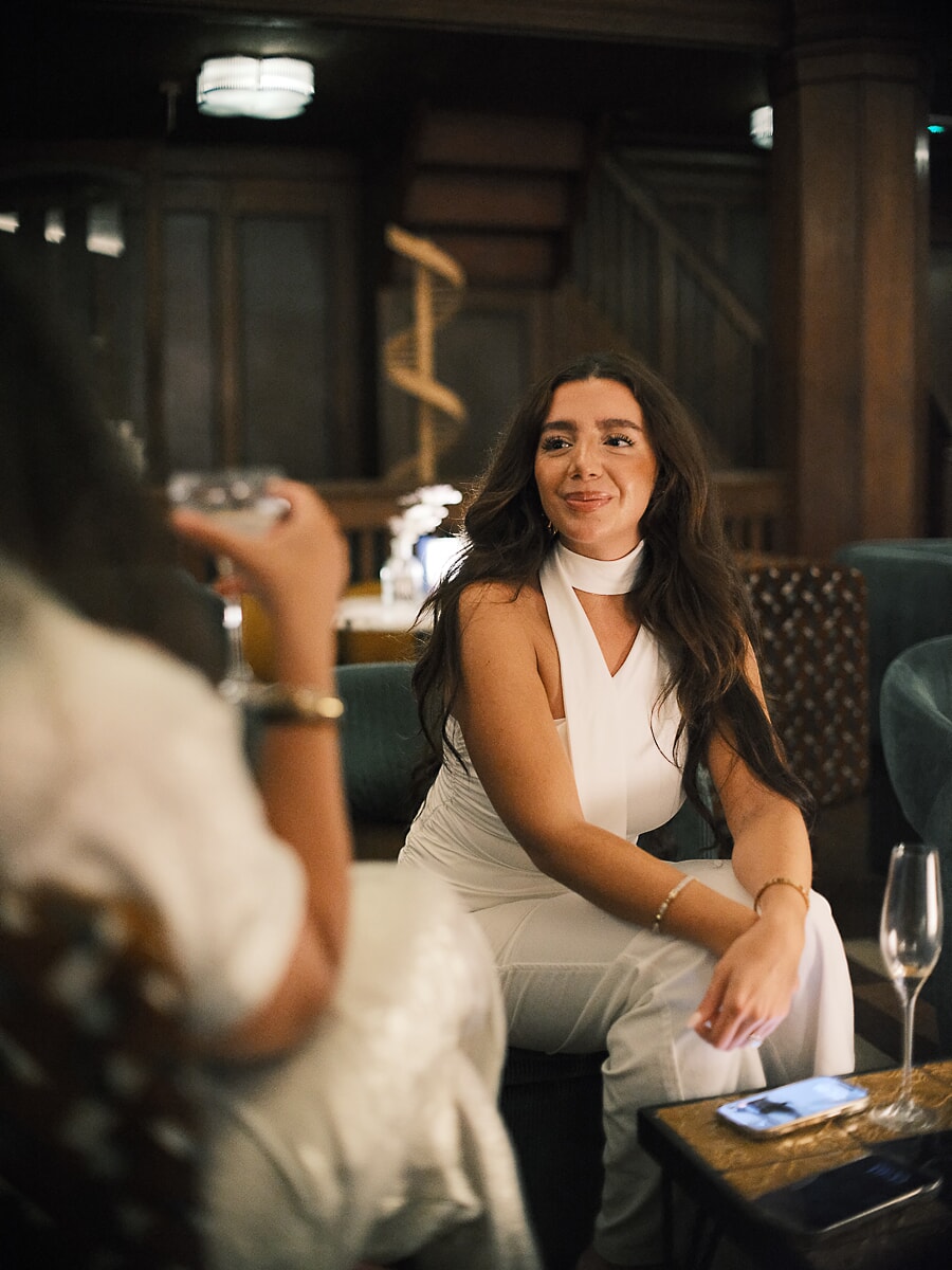 Guest in a white halter jumpsuit smiling in a wood-panelled lounge with green velvet sofas, business event photography in Amsterdam