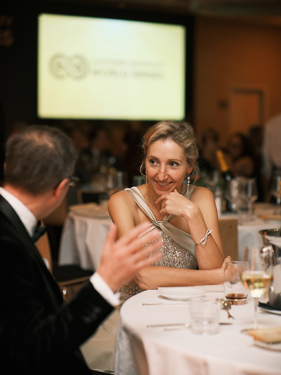Guest in a sequinned gown listening to a man in black tie across a gala table, the screen glowing behind, an evening business event in Amsterdam