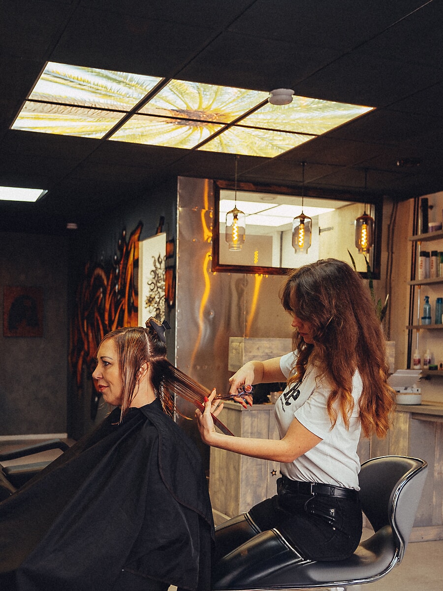 Hairdresser sectioning a client's wet hair in a salon with a printed light-panel ceiling, branding photography for an Amsterdam business