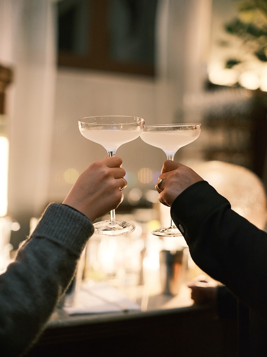 Two hands clinking coupe glasses in warm low light, candles blurred behind them