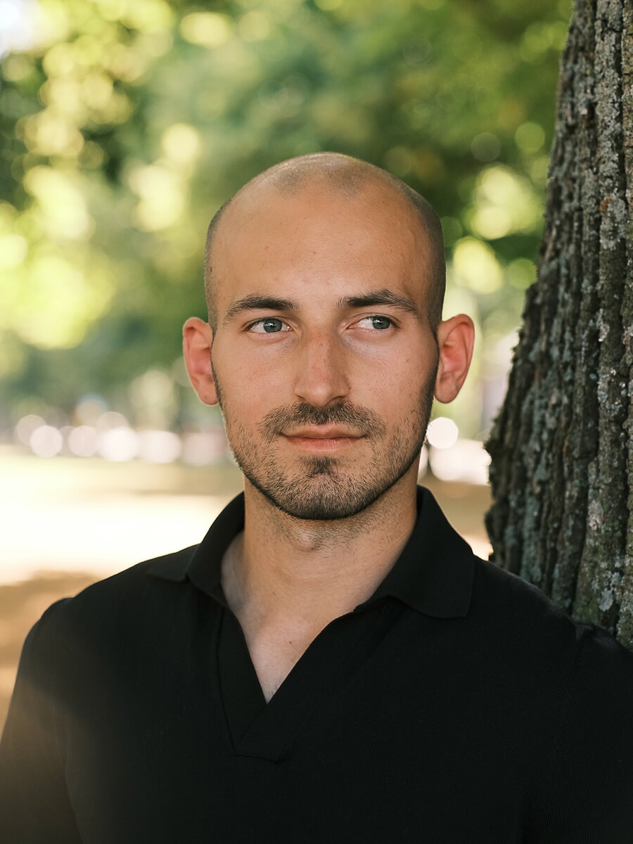 Head-and-shoulders portrait of a man in a black polo beside a tree trunk, green summer bokeh behind him