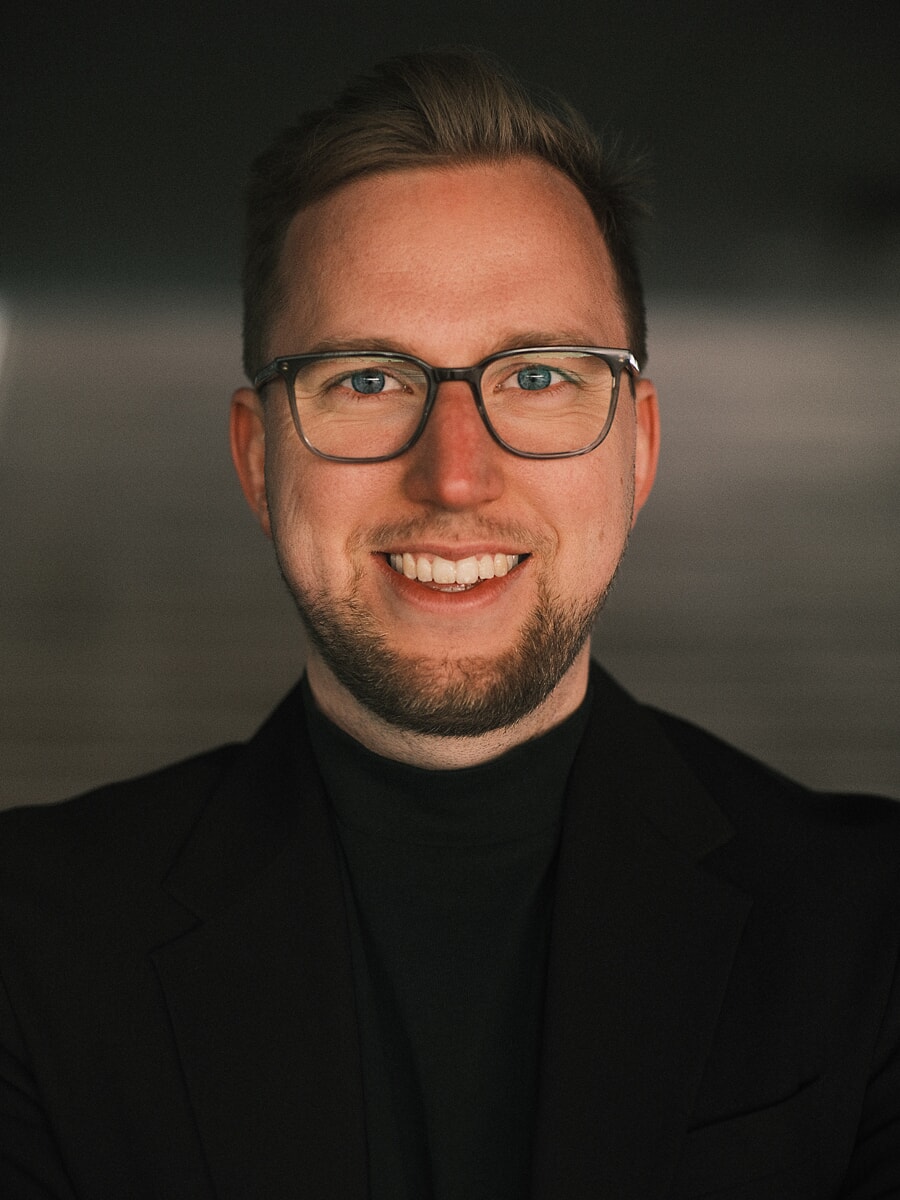 Close headshot of a man in glasses and a black blazer over a rollneck against a dark background