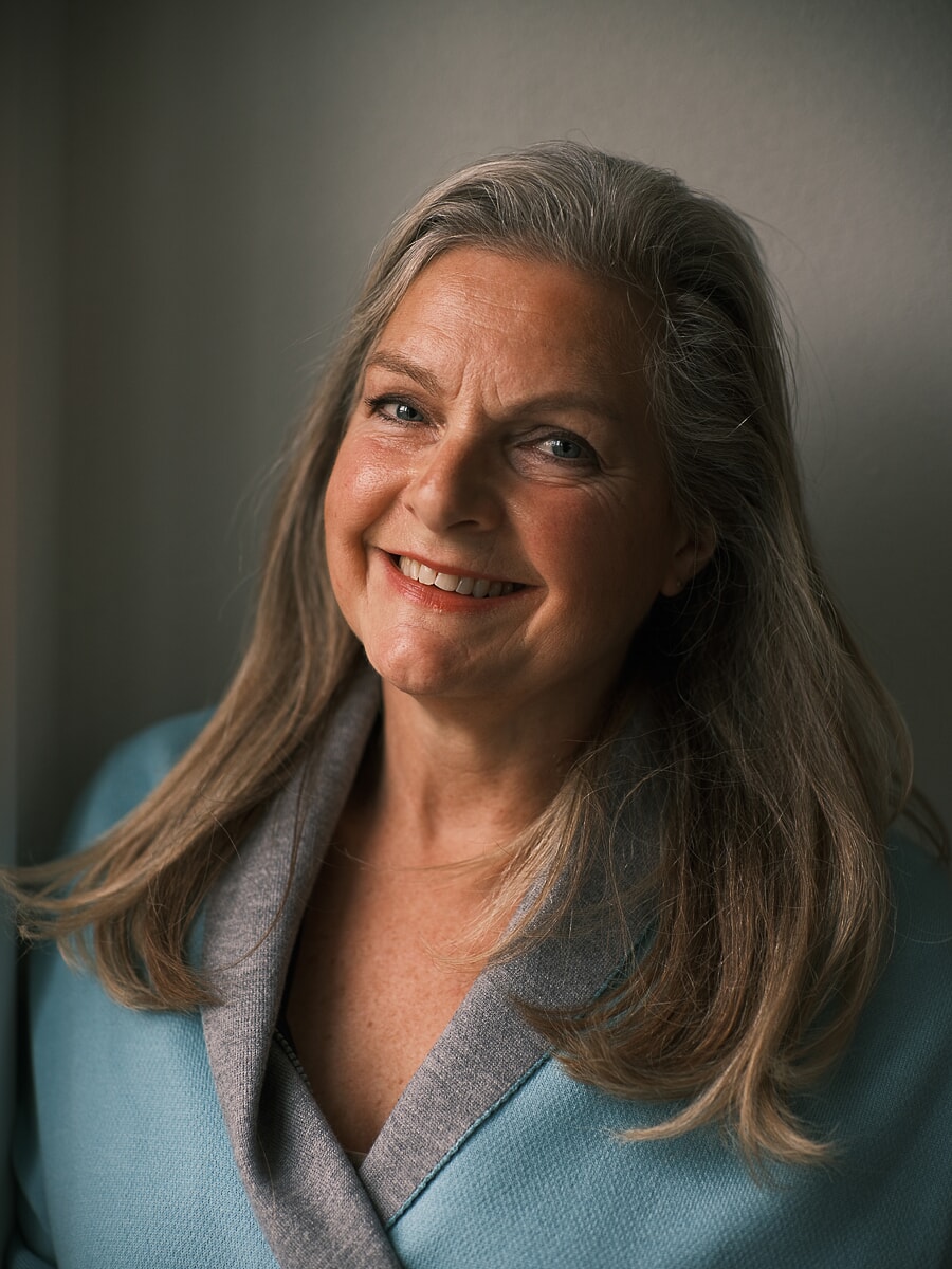 Woman with long grey-blonde hair in a pale blue jacket turned into the window light, a business headshot in Amsterdam