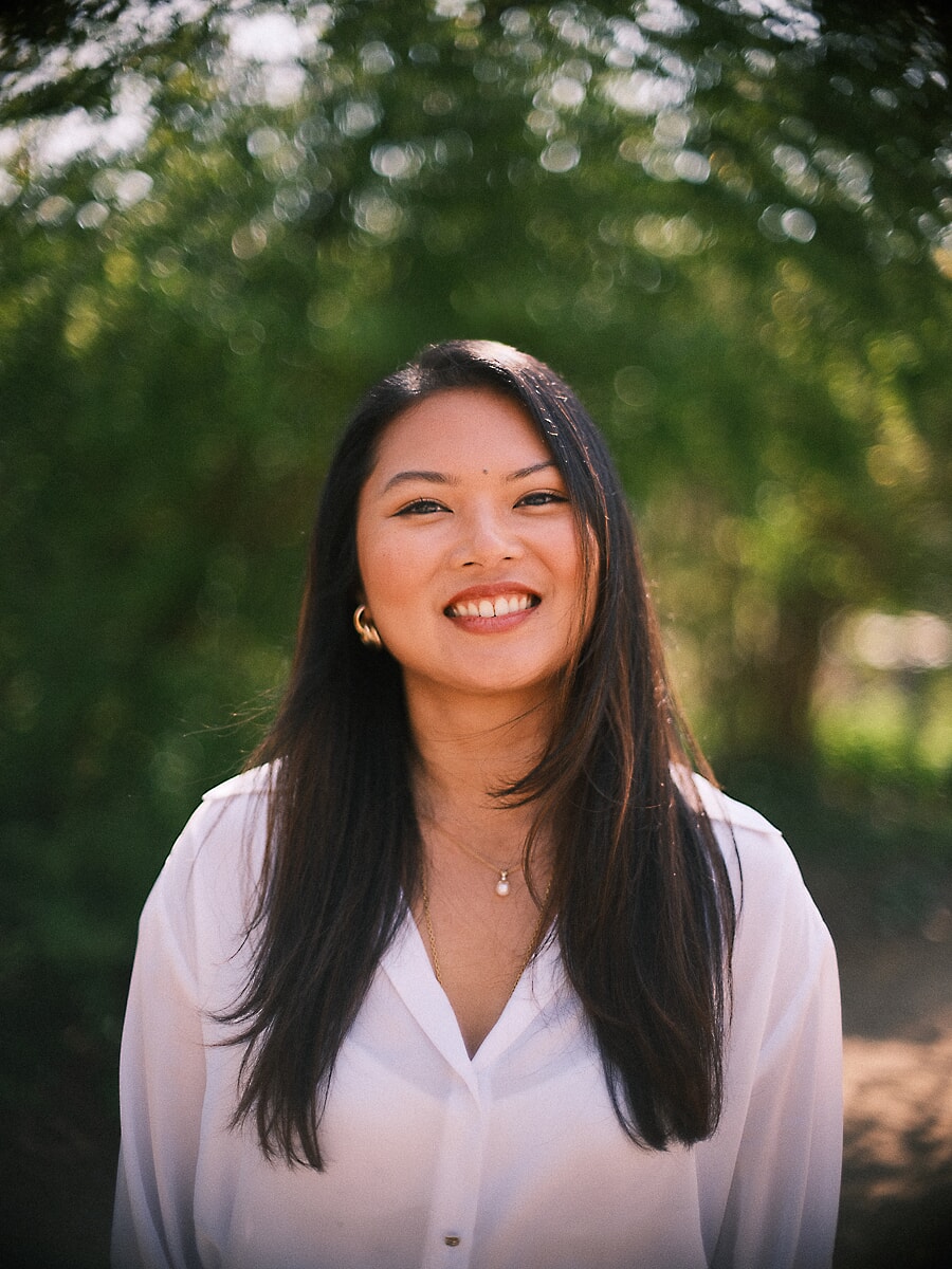Head-and-shoulders portrait of a woman in a white blouse in a park, backlit green trees behind her