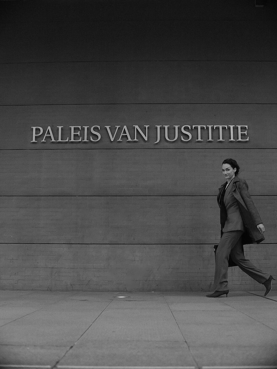 Lawyer in a suit and coat striding past the carved lettering of a Dutch courthouse, a wide black-and-white business portrait