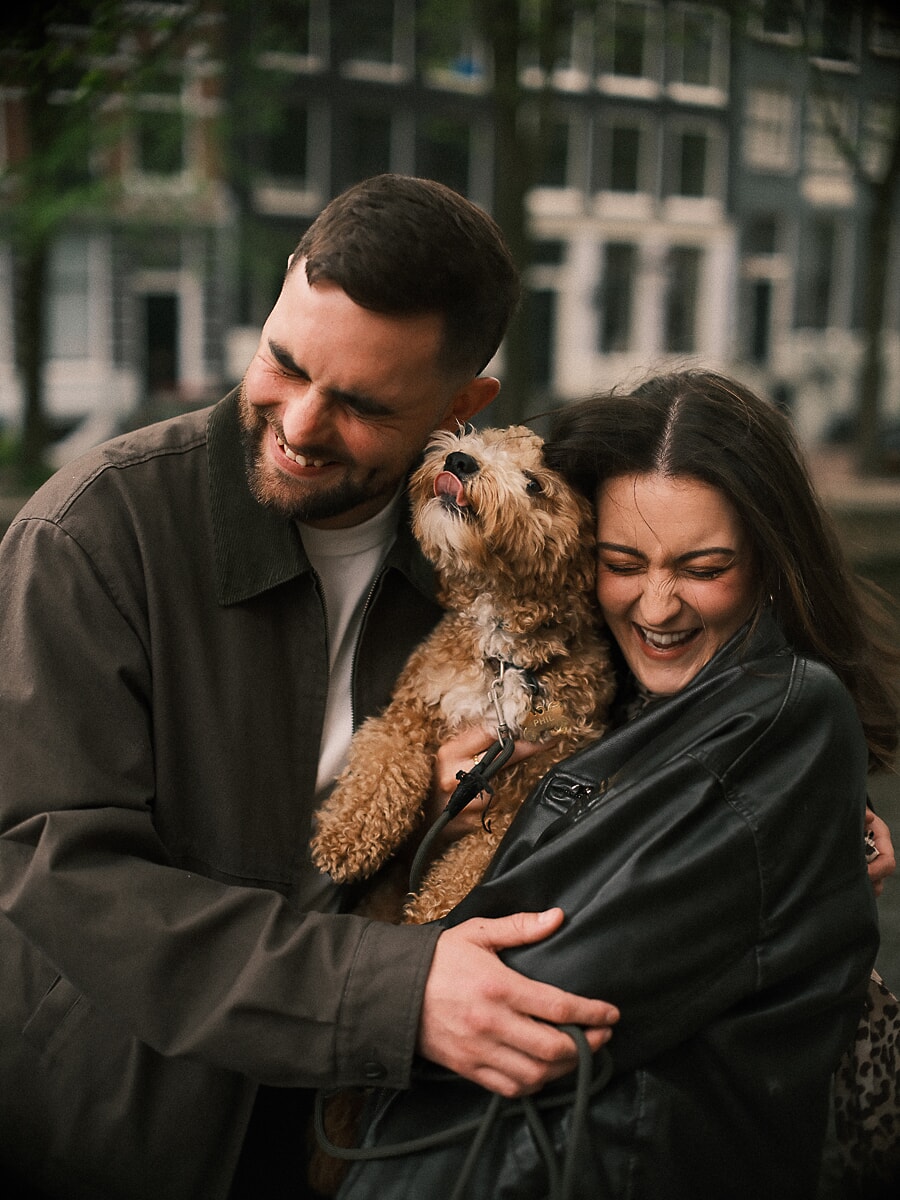 Couple laughing as they hold their small dog on an Amsterdam canal street during their loveshoot