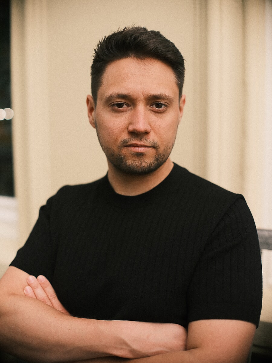 Close portrait of a man with his arms crossed in a black ribbed tee, looking straight into the lens