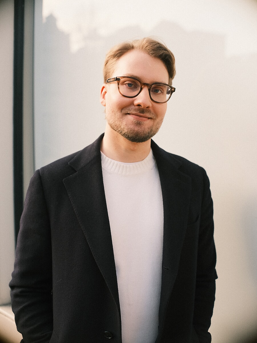 Head-and-shoulders portrait of a man in glasses and a black coat over a white knit against a plain pale wall, for an online dating profile