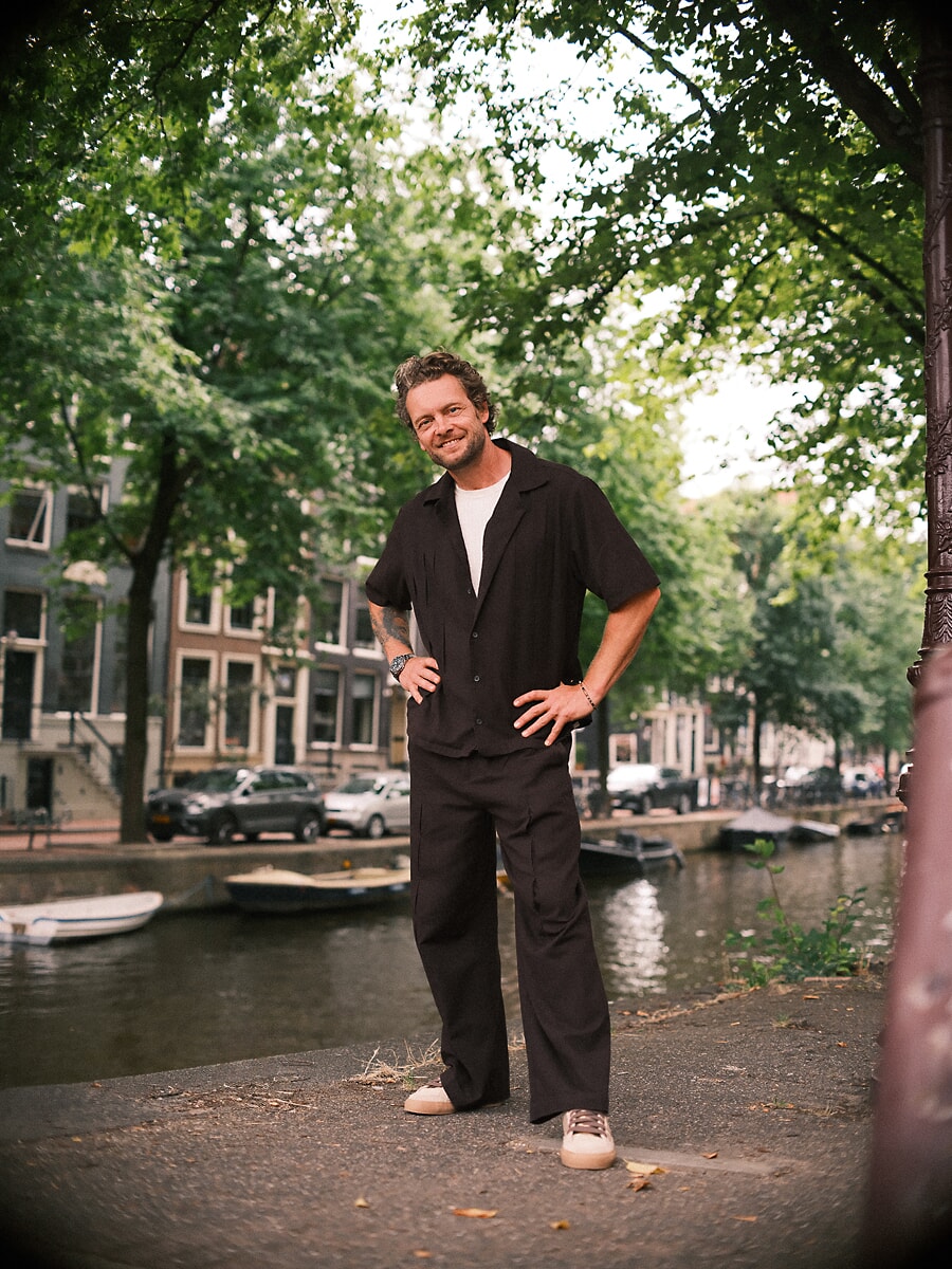 Man standing full length with a hand on his hip in a black shirt and wide trousers beside an Amsterdam canal, a relaxed confidence portrait