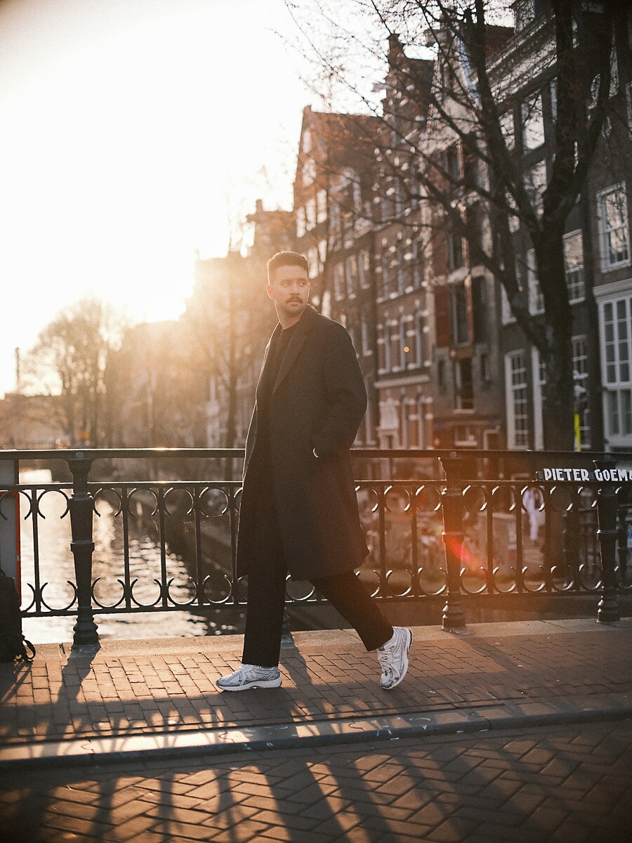 Man in a long black coat crossing a bridge in low winter sun, the railing and canal houses in silhouette behind him