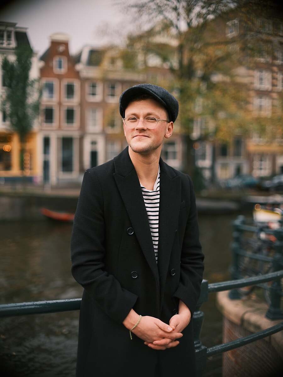Man in a flat cap and round glasses with his hands clasped, a black coat over a striped tee, a portrait beside an Amsterdam canal