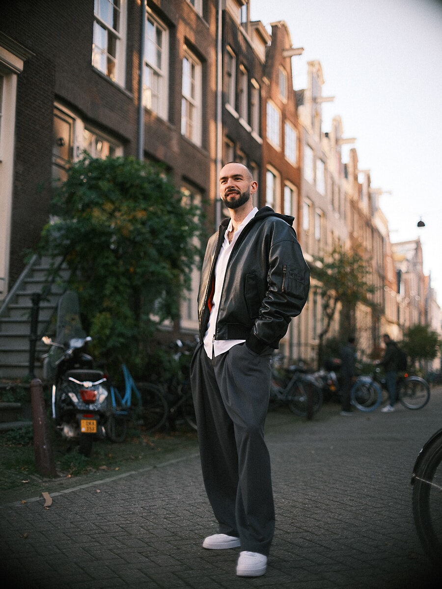 Man in a black leather bomber and wide grey trousers standing in a canal street in low autumn sun, a full-length portrait in Amsterdam