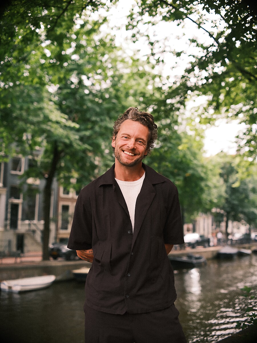 Man with curly hair laughing in a black camp shirt beside a canal full of moored boats, a summer portrait in Amsterdam