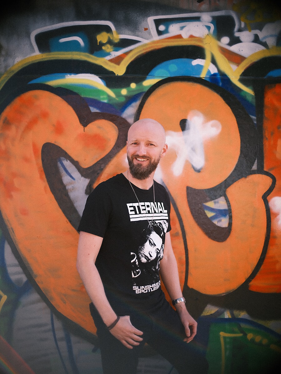 Man with a beard laughing in front of a large orange graffiti piece, a vivid frame from his dating photoshoot
