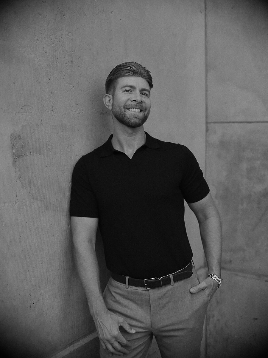 Man leaning against a plain concrete wall with one hand in his pocket, a relaxed black-and-white portrait