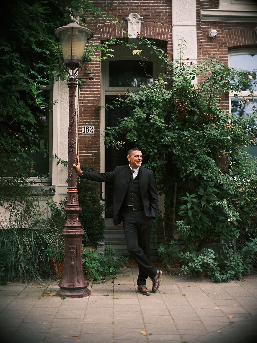 Man in a three-piece suit leaning on an ornate lamppost outside a brick house, climbing greenery around him, a portrait from his dating photoshoot in Amsterdam