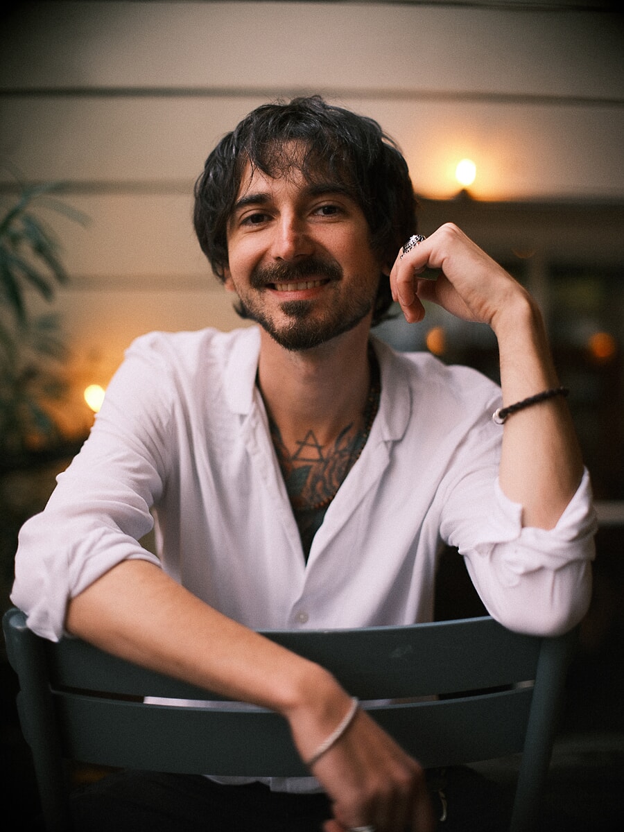 Man in an open white shirt sitting on a cafe terrace at night, warm lights blurred behind him, an evening portrait from a dating shoot in Amsterdam