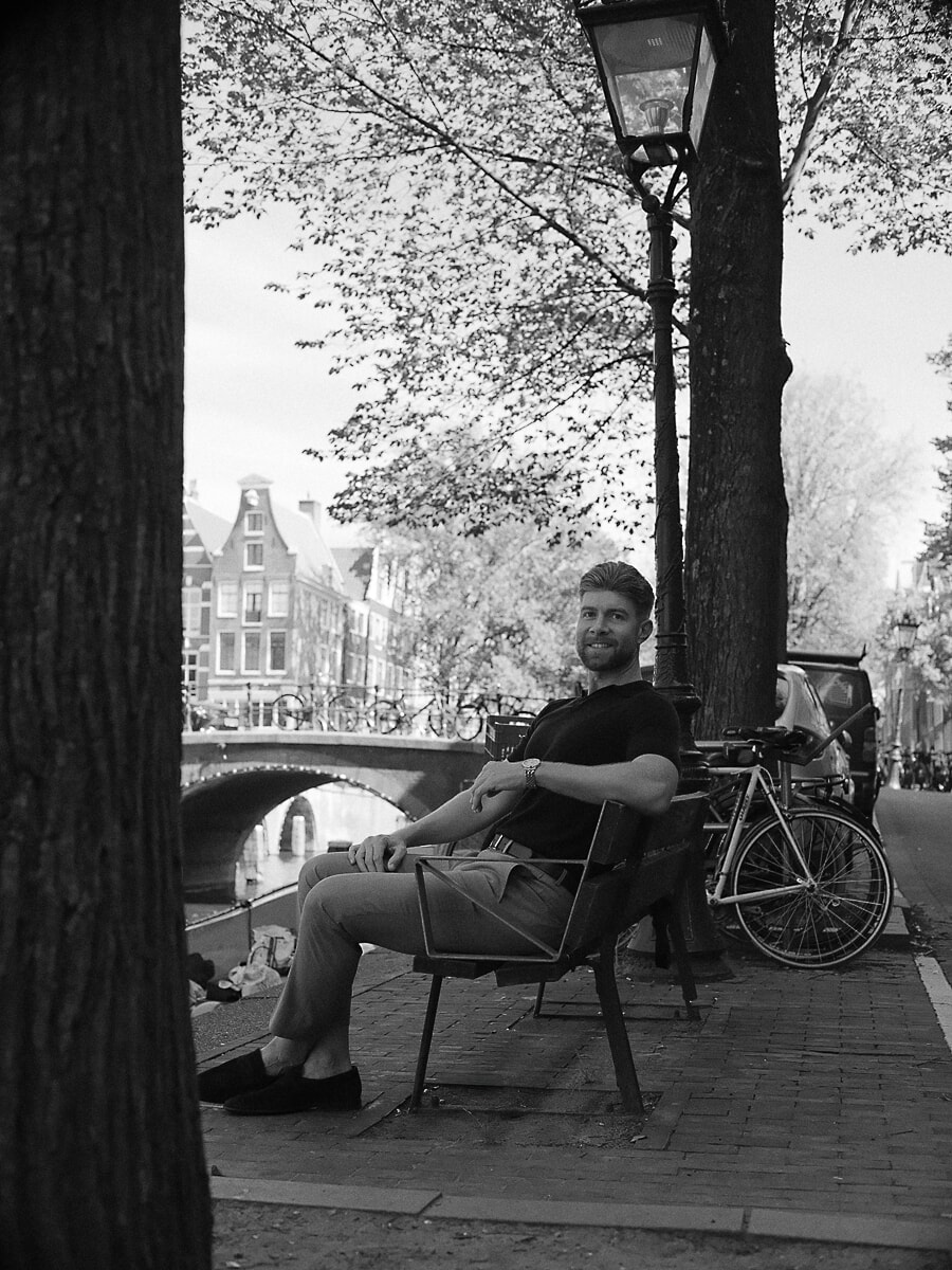 Man in a dark polo sitting on a bench beneath the trees along an Amsterdam canal, a black-and-white portrait