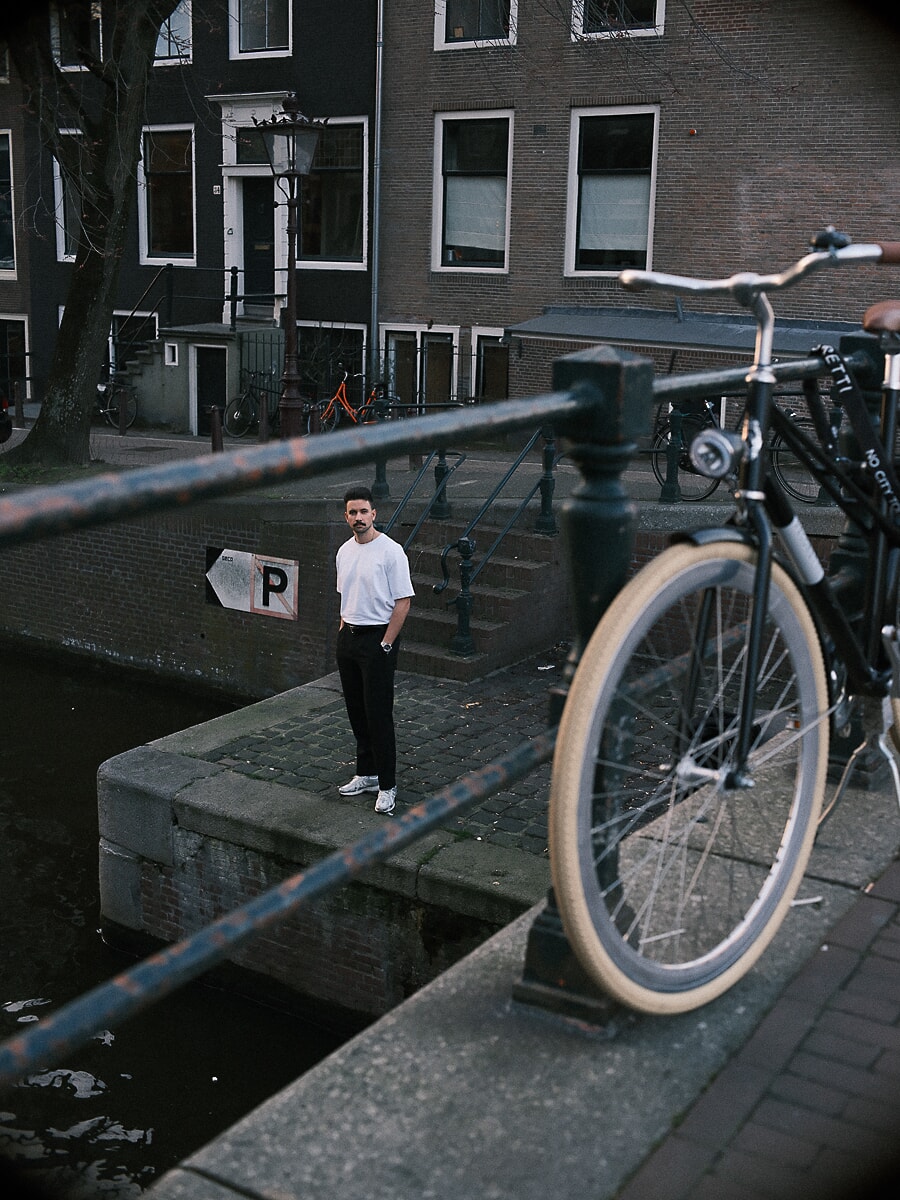 Man in a white tee standing on a canal quay, seen past a bicycle wheel and railing, a documentary portrait in Amsterdam