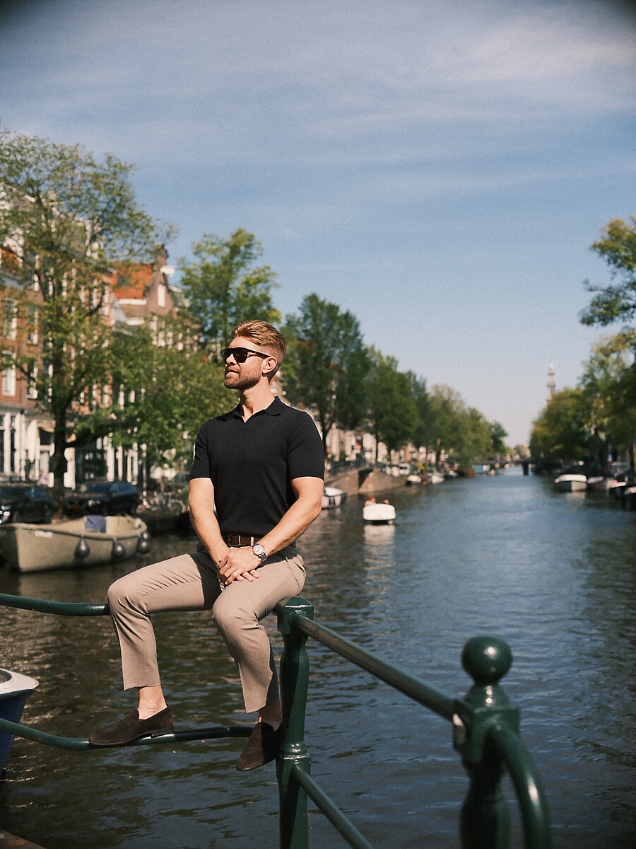 Man in sunglasses perched on a green canal railing with a wide view down the water, a lifestyle portrait in Amsterdam