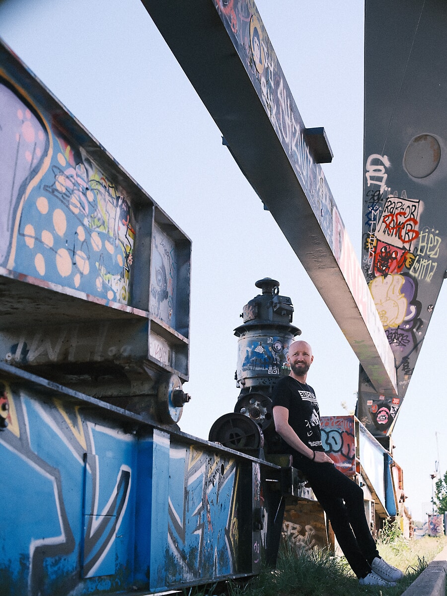 Man seated on graffiti-covered steel beams beneath a bridge structure against a pale sky