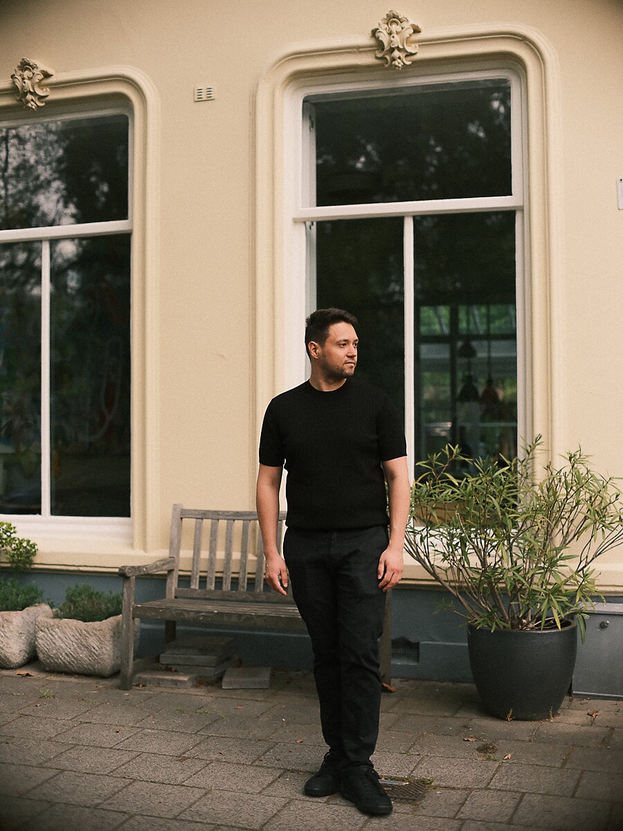 Man in a black tee standing before a cream villa with tall ornate windows, a bench and potted shrub beside him