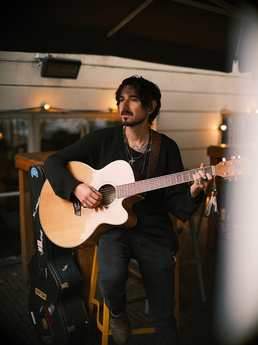 Man playing an acoustic guitar on a covered terrace at dusk, his case open beside him under warm lamps