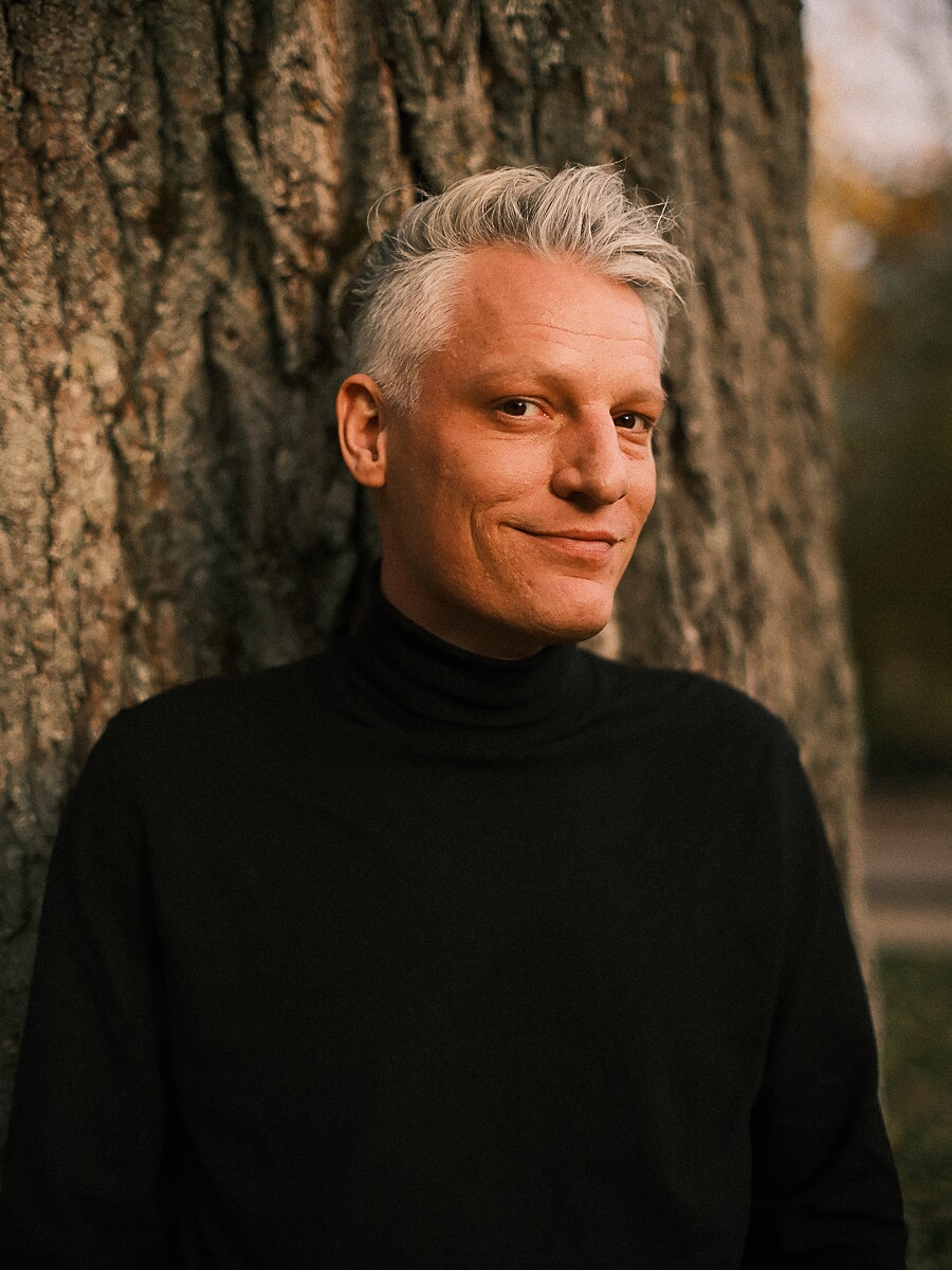 Man with grey hair in a black rollneck standing against a broad tree trunk in warm autumn light, a natural portrait