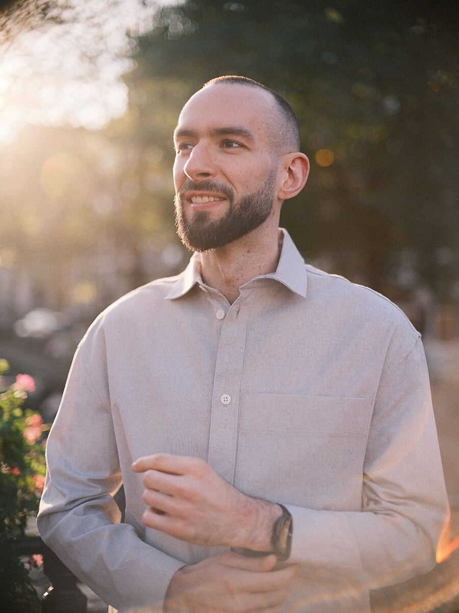 Close portrait of a bearded man in a pale linen shirt, strong sun flare washing in from the left past pink flowers