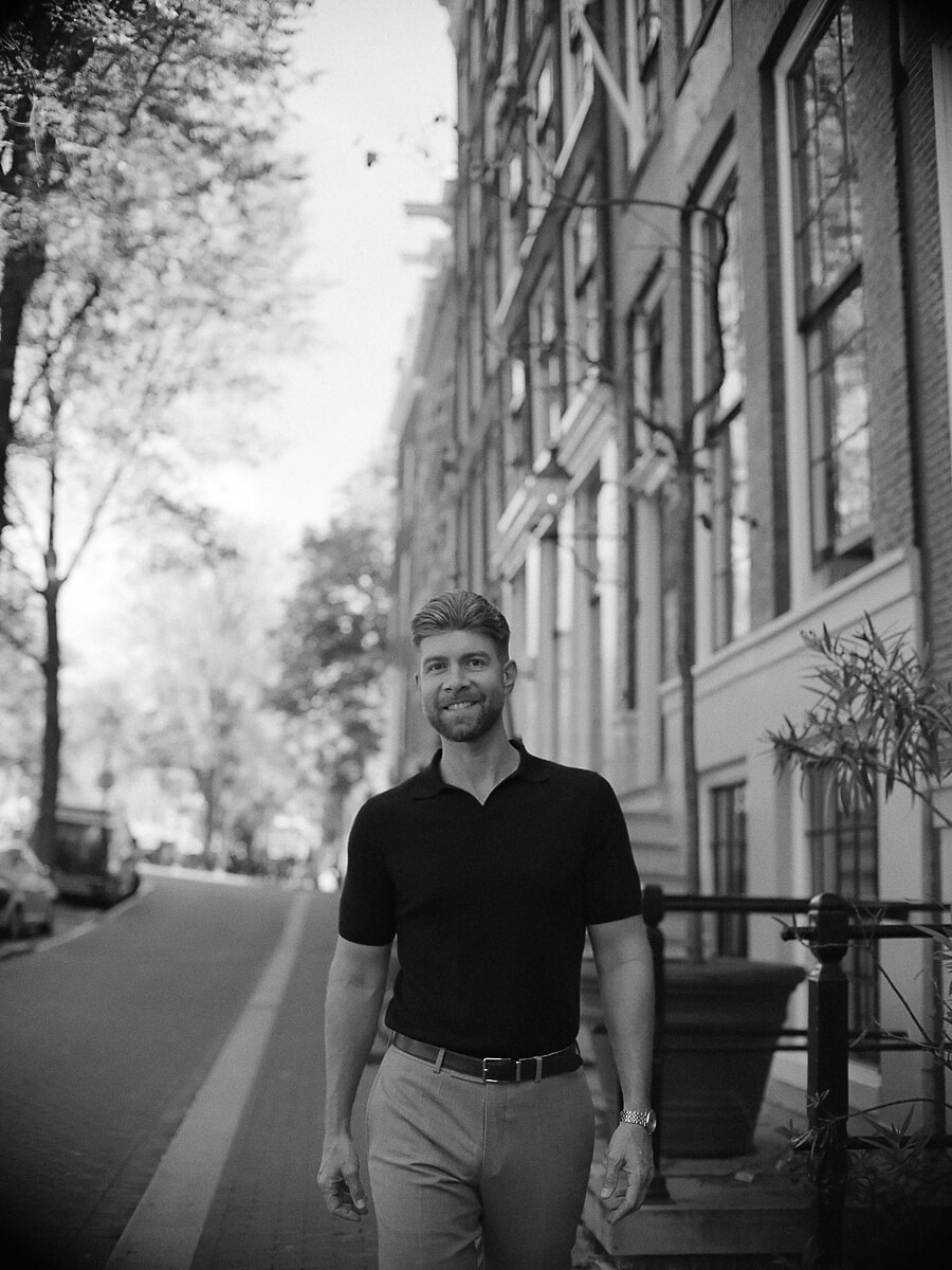 Man in a dark polo walking a leafy canal street, a wide black-and-white frame from his portrait session