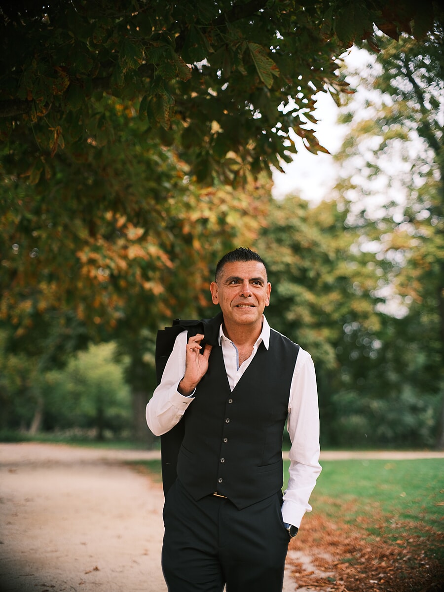 Man in a waistcoat walking a park path with his jacket over one shoulder, chestnut leaves overhead, an unhurried portrait from his dating shoot in Amsterdam
