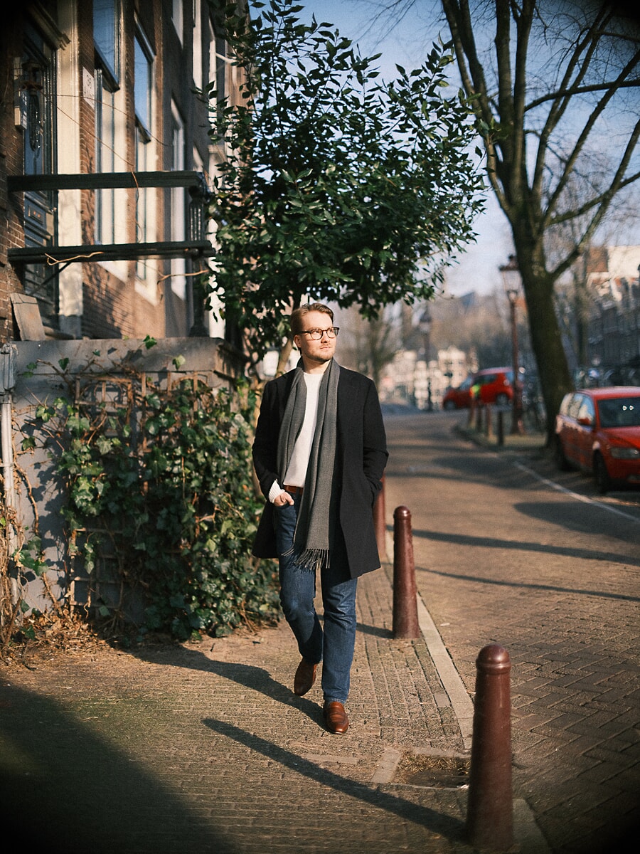 Man in glasses and a long black coat walking a sunlit canal street in winter, a candid dating portrait in Amsterdam