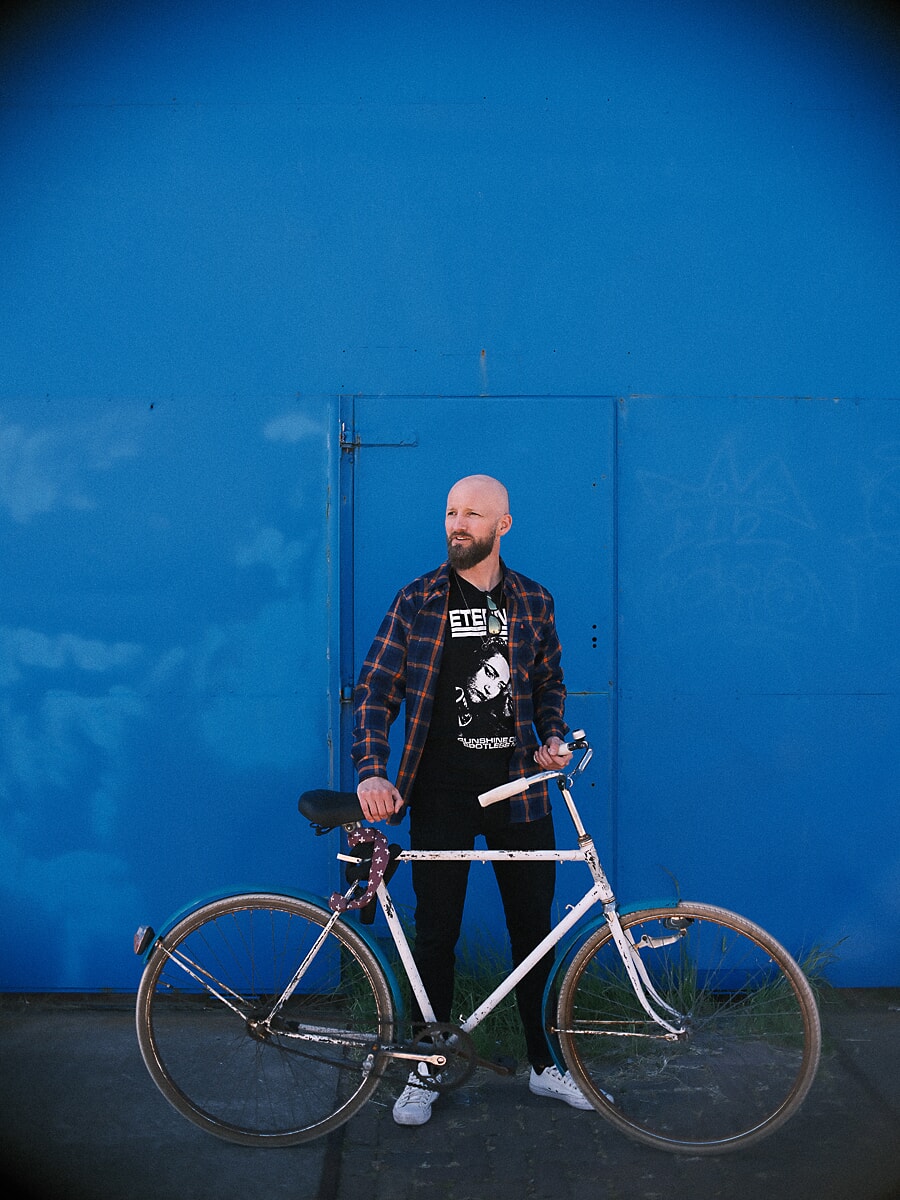 Man in a plaid shirt standing behind a white vintage bicycle against a bright blue wall, the paint filling the whole frame behind him