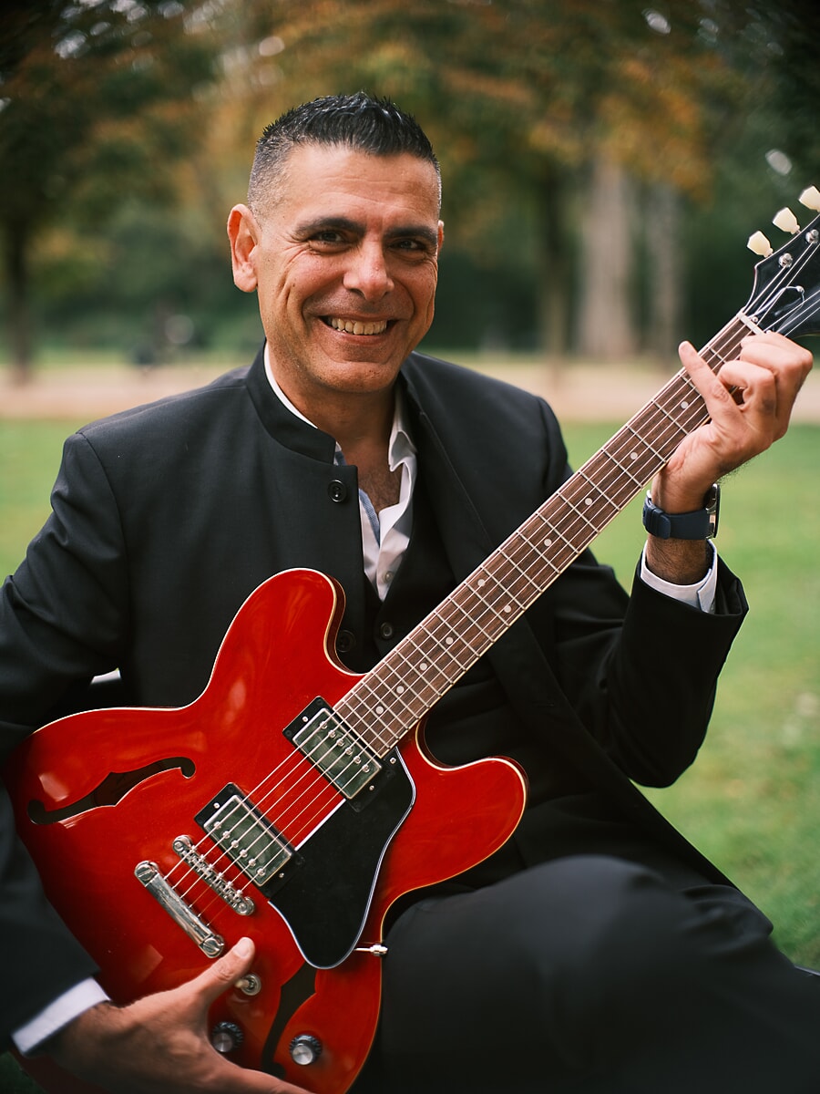 Man in a black suit sitting on the grass with a red semi-hollow guitar, laughing toward the camera, a portrait from a dating session in Amsterdam