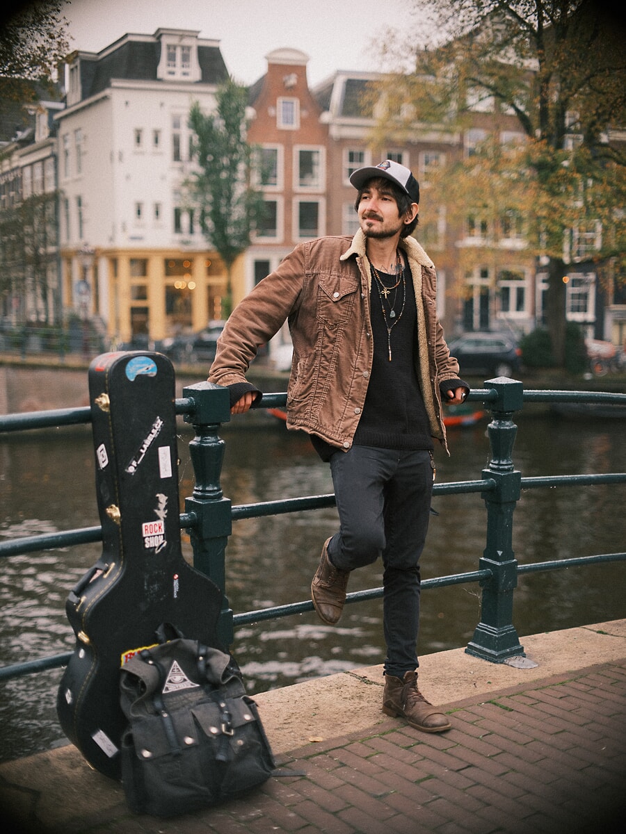 Man in a cap and corduroy jacket leaning on a canal bridge railing with his guitar case beside him, a dating portrait in Amsterdam