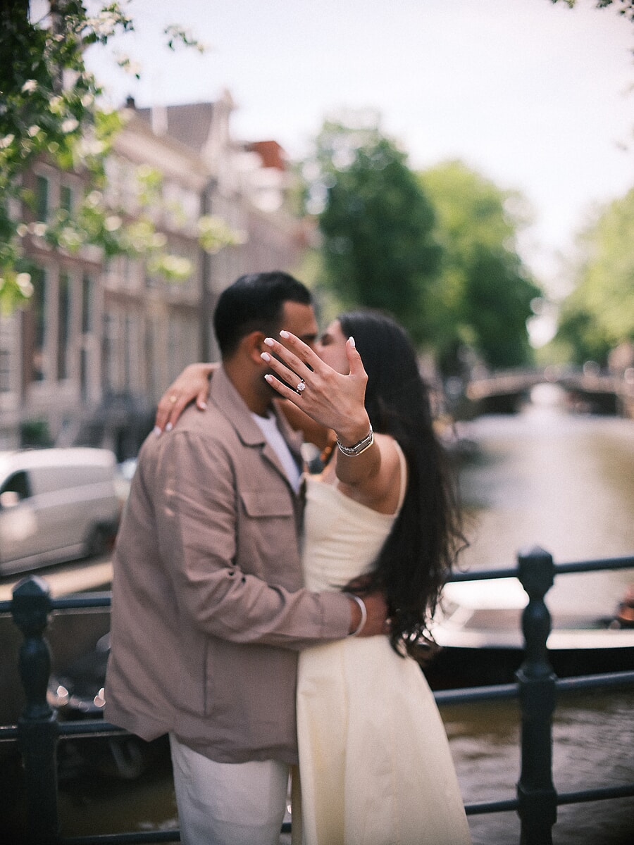 Newly engaged couple kissing behind her raised hand, the engagement ring catching the light on a canal bridge