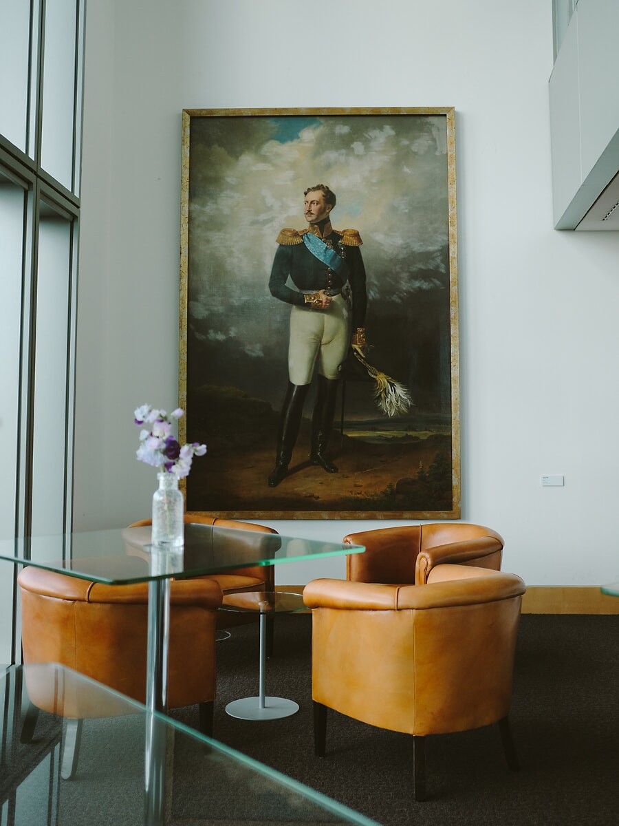 Office lounge corner with tan leather tub chairs and glass tables beneath a large classical portrait, the interior of an Amsterdam business
