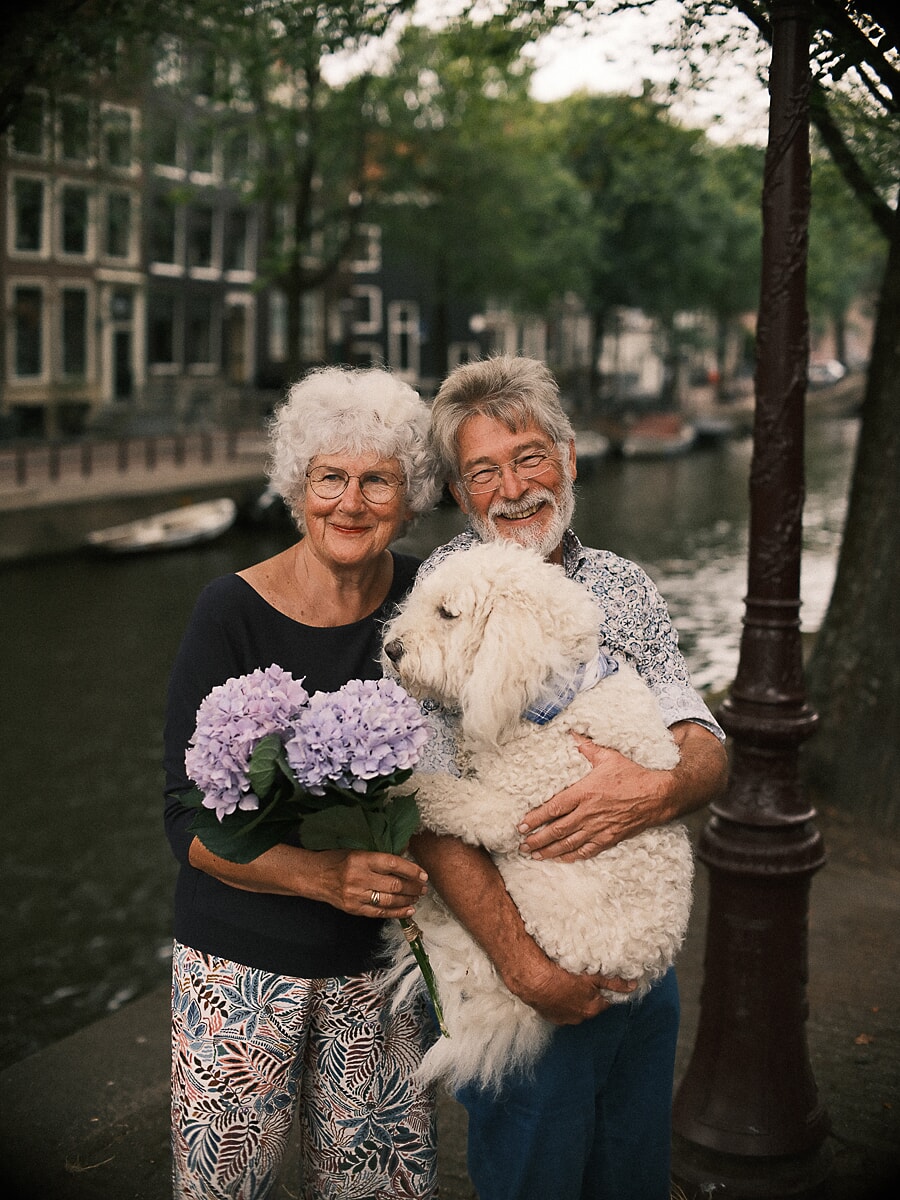 An older couple laughing on a canal quay in Amsterdam, she holds lilac hydrangeas and he carries their big curly-coated dog