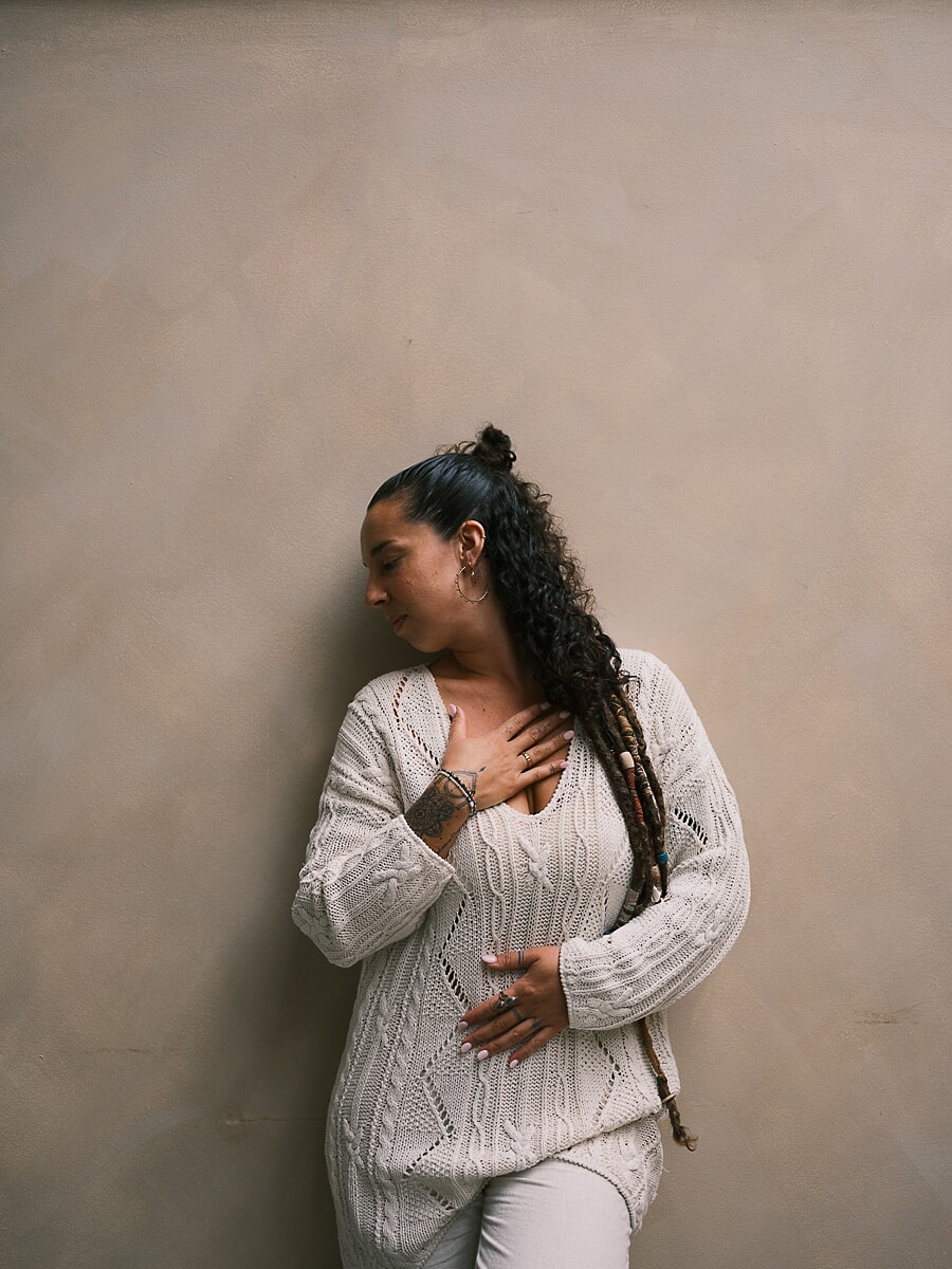 Practitioner in a cream cable-knit leaning against a plaster wall with her eyes closed and one hand on her chest