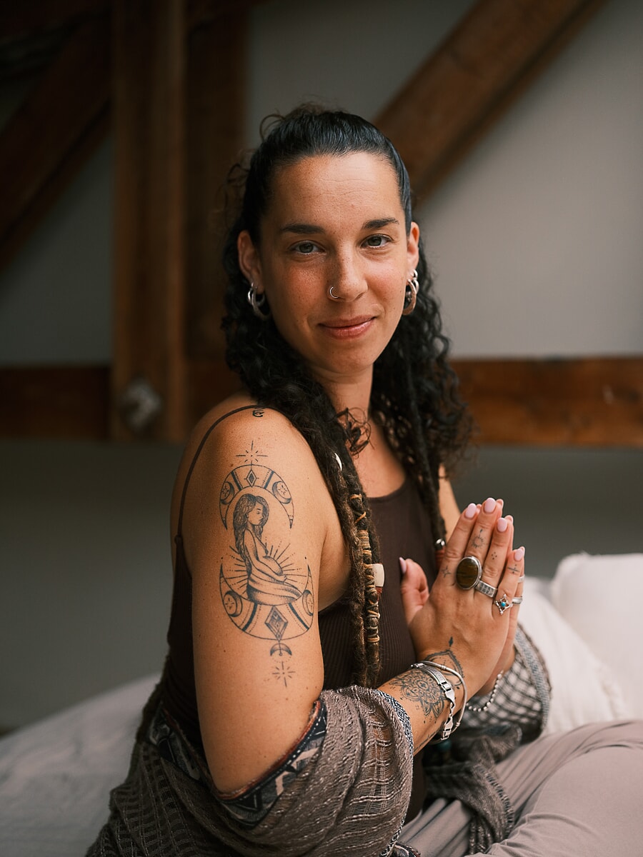 Practitioner sitting with her hands pressed together on a low bed, timber attic beams rising behind her