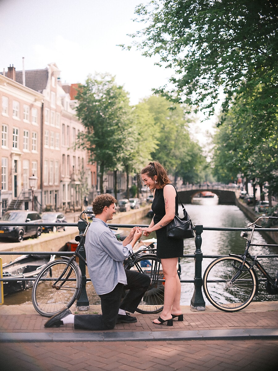 Surprise marriage proposal between parked bicycles on an Amsterdam canal bridge, his partner laughing as he kneels